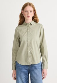 Light olive green button-up shirt with a pointed collar, long sleeves, and rounded hem. Soft texture, with visible front button placket and cuff details.