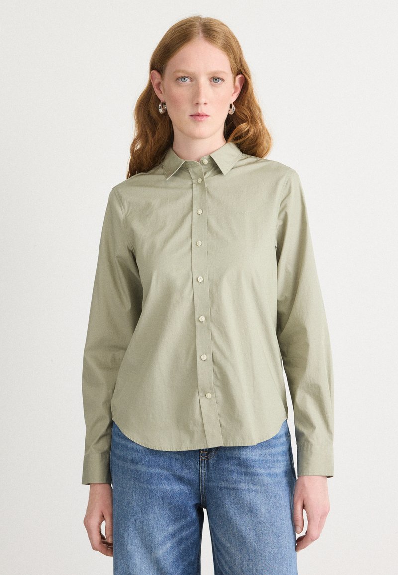 Light olive green button-up shirt with a pointed collar, long sleeves, and rounded hem. Soft texture, with visible front button placket and cuff details.