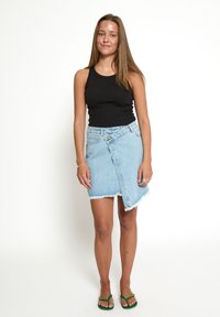 Gonna avvolgente in denim azzurro chiaro con orlo sfilacciato, vita alta e chiusura con bottoni laterali. Abbinata a una canotta nera e ciabatte verdi.