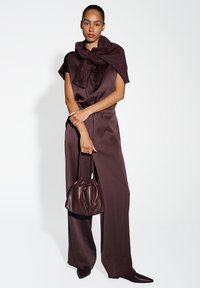 Burgundisches Outfit mit einer locker sitzenden, knöpfbaren Bluse und einer weit geschnittenen Hose. Abgerundet mit einer passenden Tasche und einem strukturierten Schal, der über die Schultern drapiert ist.