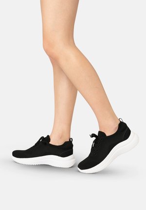 Benen met zwarte gebreide vetersneakers met dikke witte zolen op een lichtgrijze achtergrond, één voet iets opgeheven.