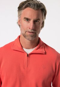 Korálový zip-up pulovr z žebrované tkaniny, s malým logem na hrudi, vysokým límcem a texturovaným, hladkým povrchem.