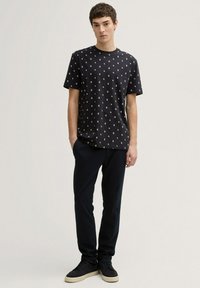 Camiseta negra de algodón con pequeño patrón floral blanco, combinada con pantalones oscuros y zapatillas negras, de pie contra un fondo neutro.