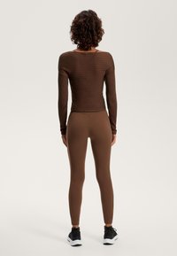 Barna bordázott, hosszú ujjú felső széles nyakkivágással, barna leggings-szel párosítva. Az öltözet sima tapintású, fekete sportcipővel kiegészítve.