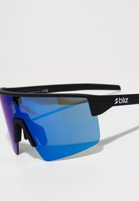 Gafas de sol con montura negra, grandes lentes azules que degradan a púrpura, diseño aerodinámico y distintivos acentos laterales. Logo de la marca visible.