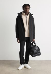 Un long manteau noir par-dessus une polaire beige à zip, associé à un pantalon noir et des baskets blanches. Tient un sac noir au design arrondi.
