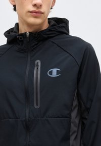 Νεαρό άτομο που φοράει μαύρο μπουφάν με κουκούλα, εμπρός φερμουάρ και κάθετη τσέπη στο στήθος με φερμουάρ, που δείχνει ένα γκρι λογότυπο.