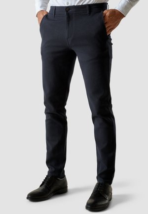 Man die nauwsluitende donkerblauwe broek draagt met zwarte nette schoenen en een wit ingestopt overhemd, staand met handen in de zakken.