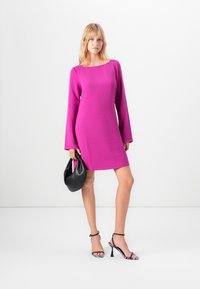 Gaun lengan panjang fuchsia dengan kerah kotak, menampilkan kain berserat. Model memegang tas hitam dan mengenakan sepatu hak tinggi hitam-putih.
