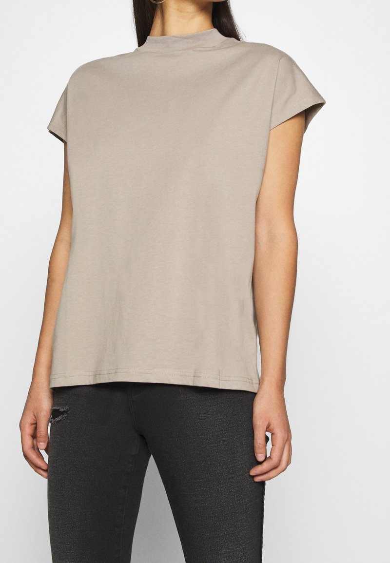 Femme portant un haut ample beige à manches courtes avec un col montant et un jean skinny noir déchiré, debout devant un fond uni.