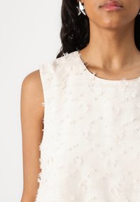 Cream-colored sleeveless top with textured, raised dots and horizontal stripes; features a round neckline and soft, lightweight fabric.
