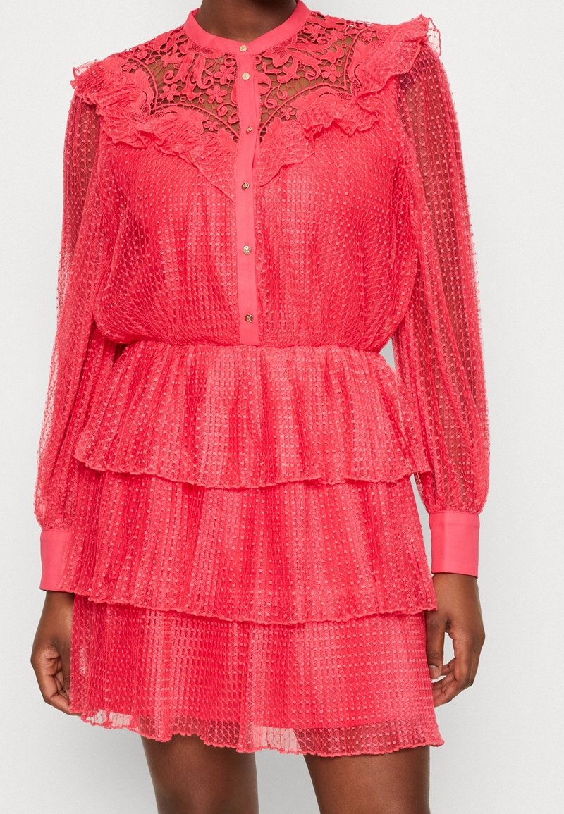 Robe rose transparente avec des volants superposés, des détails en dentelle au niveau du col, des manches longues et une fermeture à boutons, présentant un tissu texturé à pois.