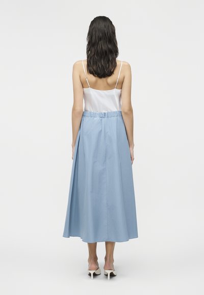 Γυναίκα με σκούρα μαλλιά, φορώντας λευκό αμάνικο τοπ και ψηλόμεση, ανοιχτό μπλε midi φούστα, όρθια με την πλάτη προς τα εμπρός σε λευκό φόντο.