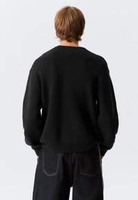 Person med kort brunt hår iført en løs svart genser og mørke jeans, sett bakfra mot en ensfarget lys bakgrunn.