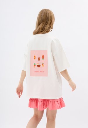 Weißes übergroßes T-Shirt mit einer pinken Grafik, die Illustrationen von Eiscreme und Desserts zeigt, kombiniert mit einem pinken Rock mit Rüschen.