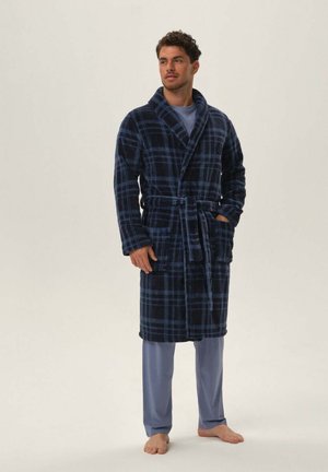 Man wearing blue plaid bathrobe tied at the waist over light blue pajamas, standing barefoot on a plain light background.