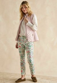 Hellrosa gesteppte Weste über einer weißen Bluse, kombiniert mit gemusterten Hosen mit floralen Designs in Grün, Rosa und Blau. Braune Mokassins.