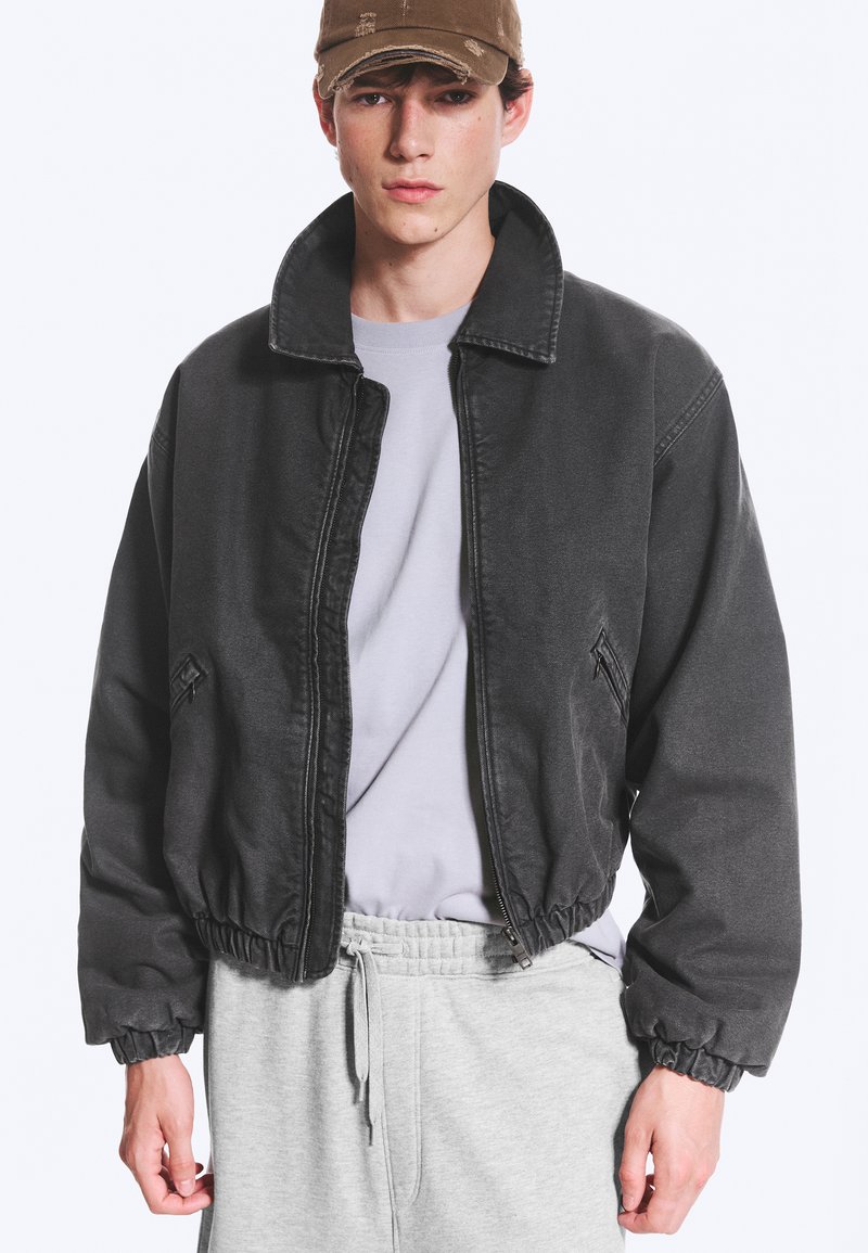 Jeune homme portant une casquette marron usée, une veste noire zippée avec des poignets élastiques, une chemise gris clair et un pantalon de survêtement gris, debout devant un fond blanc.