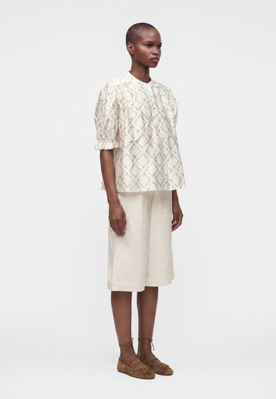 Femme portant un chemisier blanc à motifs avec des manches bouffantes, des culottes blanches jusqu'aux genoux et des ballerines marron à lacets, debout devant un fond blanc.