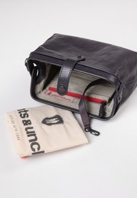 Schwarze Lederhandtasche mit strukturierter Form, rotem Reißverschlussakzent im Innenfutter und einem beigen Stoffbeutel mit schwarzem Logo und Text.