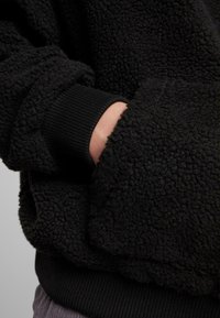 Hoodie en polaire noire présentant un extérieur texturé et doux, avec des poignets côtelés et des poches intégrées dans le design pour une fonctionnalité accrue.