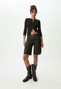 Schwarzes Langarmhemd mit Knöpfen, hoch taillierte schwarze Denim-Shorts und knielange schwarze Lederstiefel mit Schnallen-Detail.