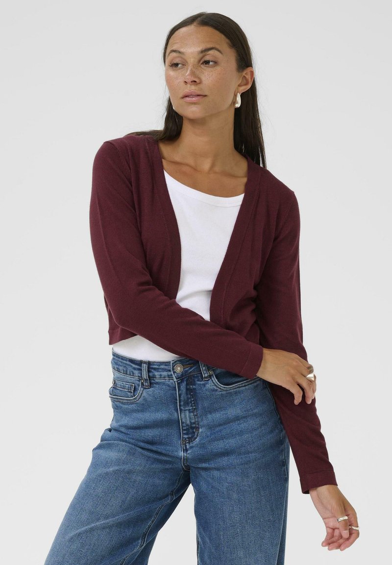Cardigan court bordeaux en tissu doux, à design ouverture devant, associé à une chemise blanche et un jean taille haute bleu.