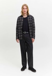 Black and white checkered flannel shirt over a black t-shirt, paired with loose black trousers and black shoes, on a neutral background.