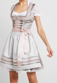 Robe traditionnelle dirndl avec un corsage décoratif argenté, des manches en dentelle et une jupe blanche imprimée, ornée d'une ceinture et de détails à ruban rose.