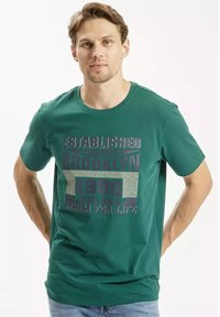 Zielona bawełniana koszulka z okrągłym dekoltem, z nadrukowanym tekstem "Założono Brooklyn 1990" oraz geometrycznymi wzorami w odcieniach szarości i bieli.