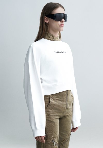Personne portant un sweat-shirt blanc surdimensionné, un pantalon marron taille haute avec des fermetures éclair et de grandes lunettes de soleil noires sur un fond gris uni.