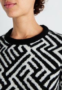 Pull en tricot noir et blanc avec un motif en zigzag audacieux, encolure ronde et tissu texturé. Gros plan sur les détails du vêtement.