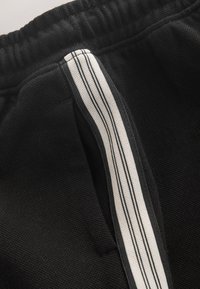 Pantalon en tissu noir texturé avec une taille élastique et une bande verticale blanche comportant deux fines lignes noires près de la poche.