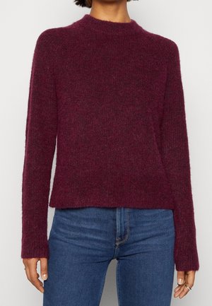 Osoba ubrana w bordowy, puszysty sweter z długim rękawem oraz niebieskie dżinsy, pokazana od połowy szyi do bioder na neutralnym tle.