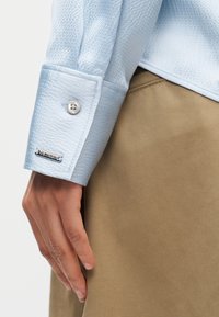 Ανοιχτό μπλε υφασμάτινο μανίκι πουκάμισου με κουμπί και μεταλλική ετικέτα Calvin Klein, το οποίο ακουμπά κοντά σε χέρι και ύφασμα μπεζ παντελονιού.