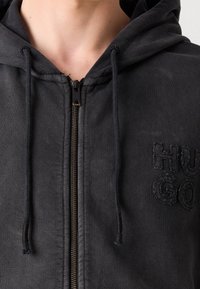 Schwarze ZIP-HOODIE aus strukturiertem Stoff, mit einer Kordelzugkapuze und gesticktem "HUGO"-Logo auf der Brust.