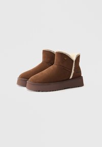 Bottines en suédine marron avec une doublure en fausse fourrure et une semelle en caoutchouc texturée. Présente des coutures contrastantes et un petit logo sur le côté.
