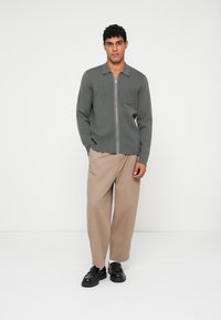 Grå zip-genser med flat krage og brystlomme, kombinert med beige vide bukser og svarte klumpete sko. Enkel design og teksturer.