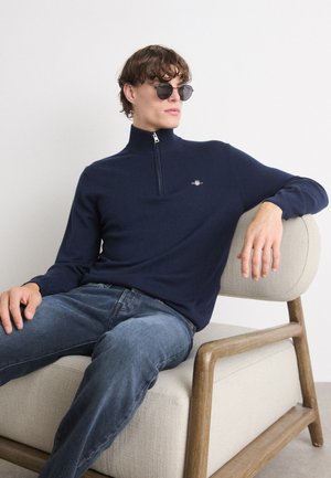 Navy zip-up sweater with long sleeves, knitted texture, featuring a small logo. Worn with blue jeans, seated on a light armchair.