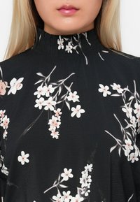 Mulher vestindo uma blusa preta de gola alta com padrões florais brancos e rosas, mostrando apenas a parte inferior do rosto e o torso superior.