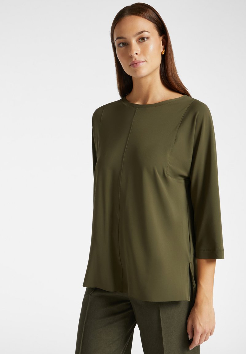 Blusa verde oliva con collo tondo, maniche a tre quarti e taglio dritto. Presenta una cucitura centrale e spacchi laterali per facilitare i movimenti.