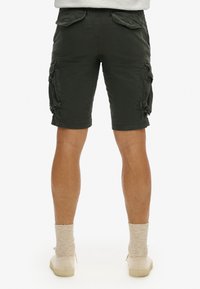 Shorts cargo en tissu vert foncé, avec des poches cargo sur les côtés, une coupe droite et des ourlets finis. Associés à des chaussettes et chaussures beiges.
