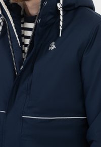 Navy waterproof jacket with a zip and snap closure, white stitching accents, and a fleece-lined hood. Features a logo on the chest.