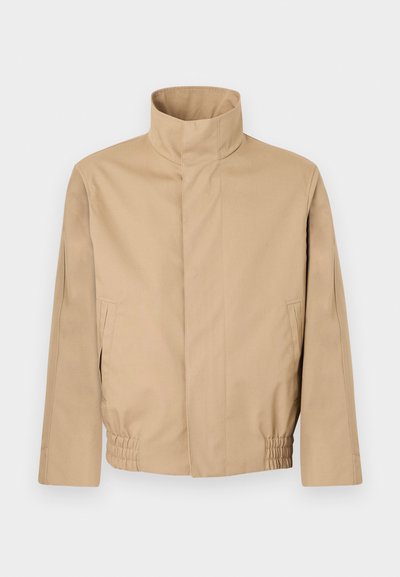 Veste beige avec col montant, coupe décontractée, ourlet élastiqué et poches latérales. Texture lisse et design minimaliste.