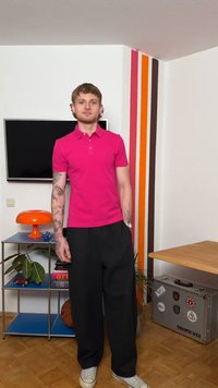 Νέος άνδρας με τατουάζ, ντυμένος με φούξια πόλο πουκάμισο και μαύρο παντελόνι, στέκεται σε δωμάτιο με τηλεόραση, πορτοκαλί λάμπα, φυτά και ριγωτή διακόσμηση τοίχου.