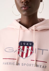 Lichtroze hoodie met koordjes en geborduurd logo met een schildontwerp in blauw en rood met de tekst "GANT AMERICAN SPORTWEAR."