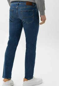 Jeans in denim blu con vestibilità slim, dotati di tasche posteriori e un'etichetta in pelle marrone. Abbinati a sneakers bianche. Texture liscia.