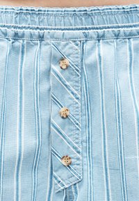 Ύφασμα με ανοιχτό μπλε και λευκές ρίγες, τρία μπεζ κουμπιά κατά μήκος του κέντρου και ελαστική μέση στο πάνω μέρος.