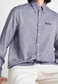 Modrá košile s dlouhým rukávem a knoflíkovým zapínáním vyrobená z texturovaného materiálu, s jemným logem "BOSS" a bílými knoflíky. V kombinaci s kalhotami z jemného manšestru světlé barvy.