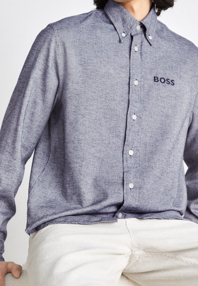 Modrá košile s dlouhým rukávem a knoflíkovým zapínáním vyrobená z texturovaného materiálu, s jemným logem "BOSS" a bílými knoflíky. V kombinaci s kalhotami z jemného manšestru světlé barvy.